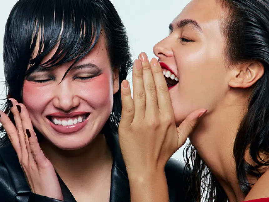 Two expressive women with wet-look hairstyles and bold beauty makeup laughing and whispering together.